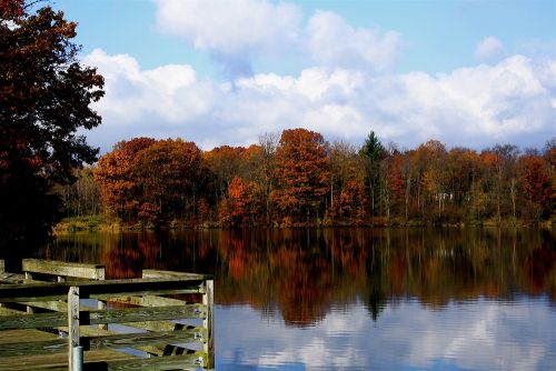 The image is of a light blue sky with fluffy white clouds in Flint Genesee. There are red trees hovering over the river. The river mirrors the blue sky and trees. 